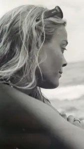 Black and white photo of a woman in profile on a beach, looking down in grief, wearing pearls.